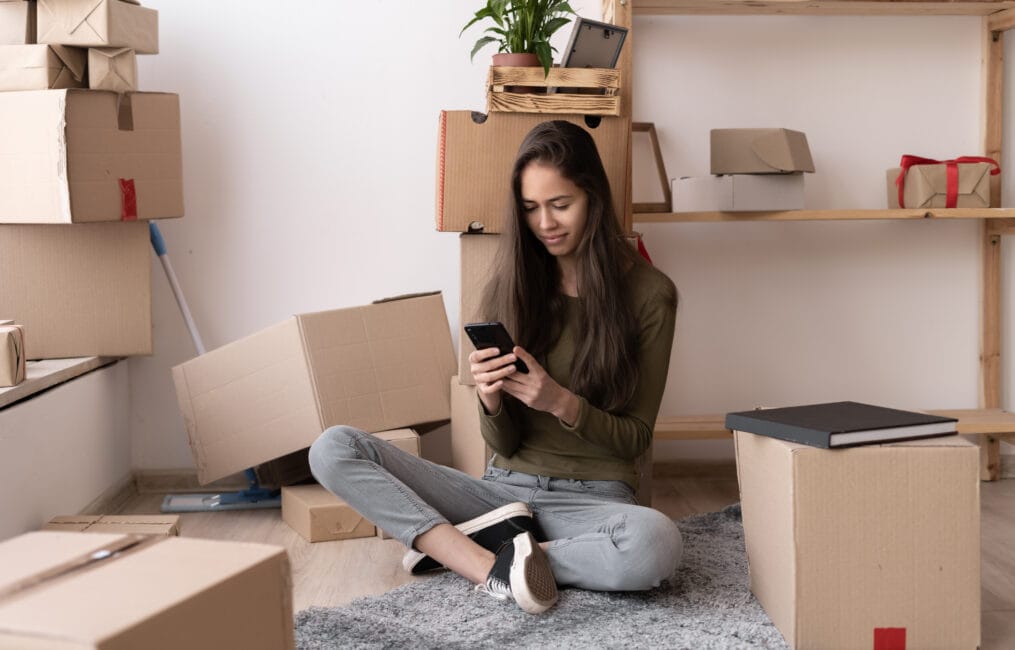 Woman using a moving app on her smartphone among cardboard boxes on moving day