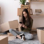Woman using a moving app on her smartphone among cardboard boxes on moving day