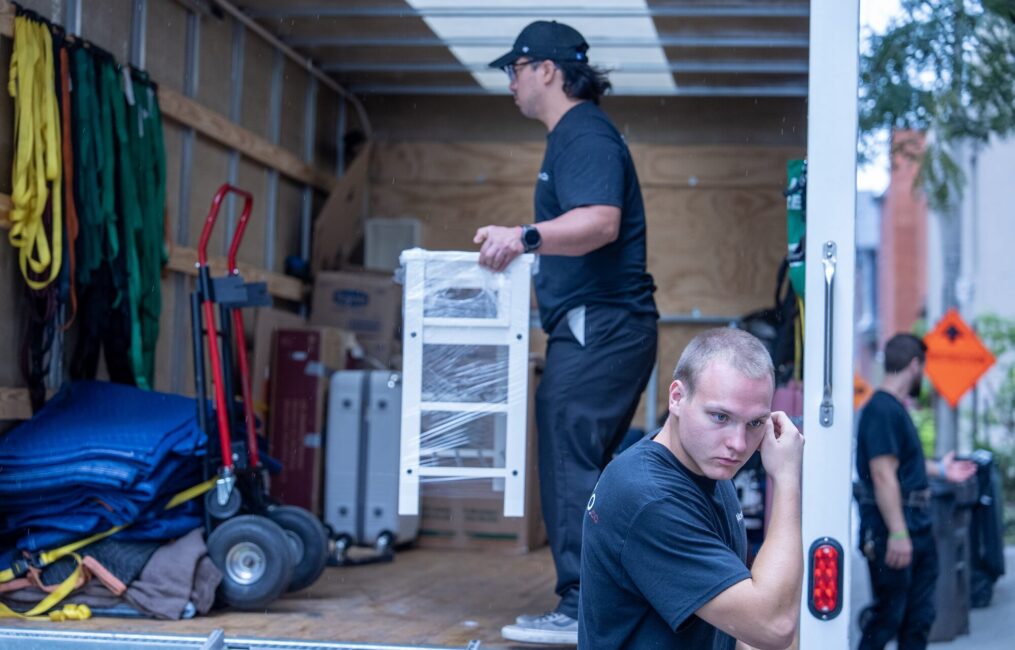 Moving Co. crew loading a white shelving unit into the truck beside dollies and moving blankets