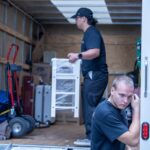 Moving Co. crew loading a white shelving unit into the truck beside dollies and moving blankets