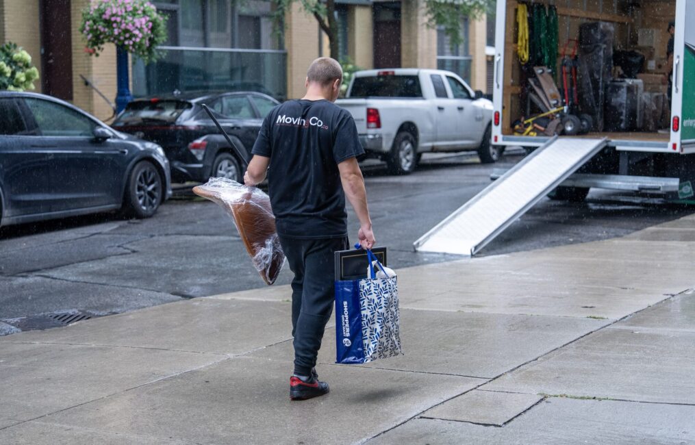 Moving Co. mover carrying a bag and a wrapped chair to the truck ramp