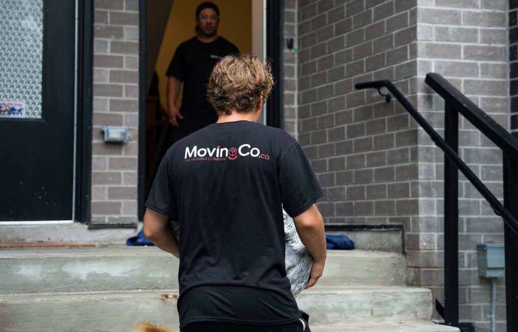 Moving Co. mover carrying a box up the front steps of a house while a teammate waits at the door
