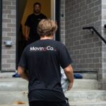 Moving Co. mover carrying a box up the front steps of a house while a teammate waits at the door