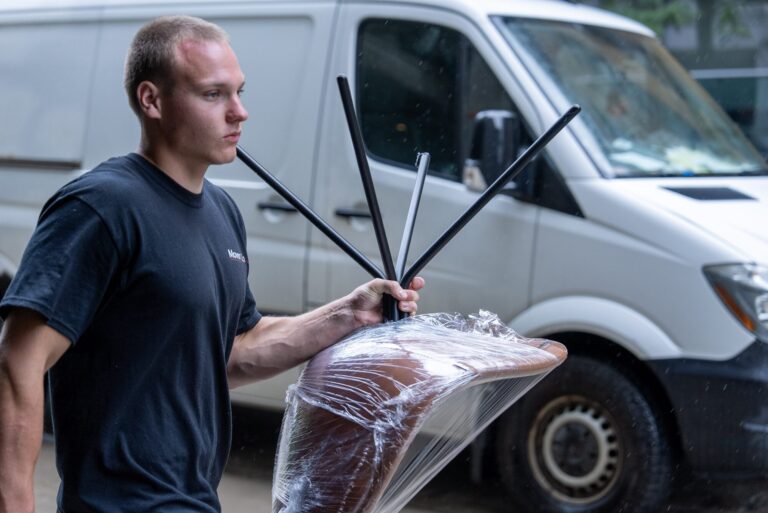 Moving Co. mover carrying a plastic-wrapped chair past a parked van