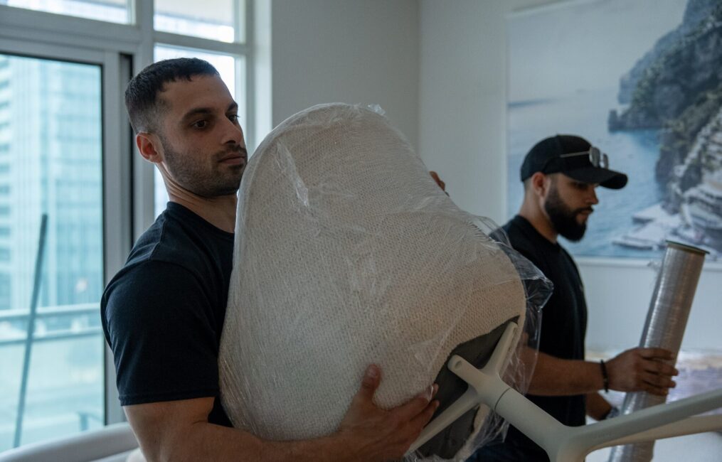 Moving Co. mover holding a shrink-wrapped chair while a teammate wraps more furniture