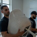 Moving Co. mover holding a shrink-wrapped chair while a teammate wraps more furniture