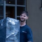 Moving Co. mover smiling while carrying a shrink-wrapped piece of furniture