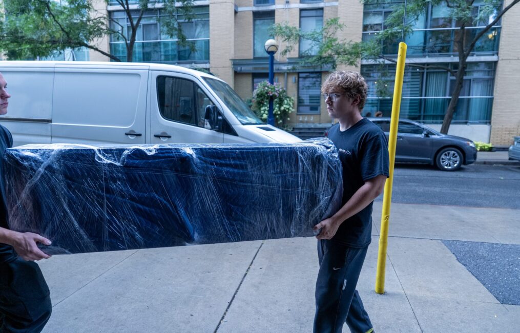 Two Moving Co. movers carrying a blanket-wrapped sofa along the sidewalk