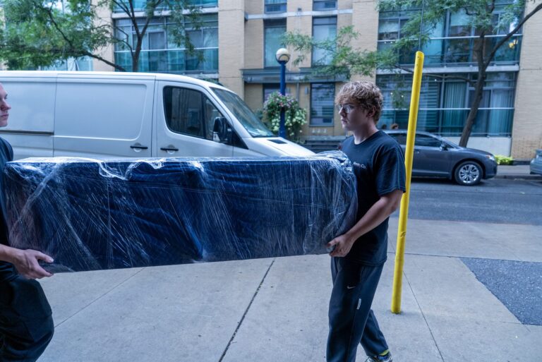 Two Moving Co. movers carrying a blanket-wrapped sofa along the sidewalk