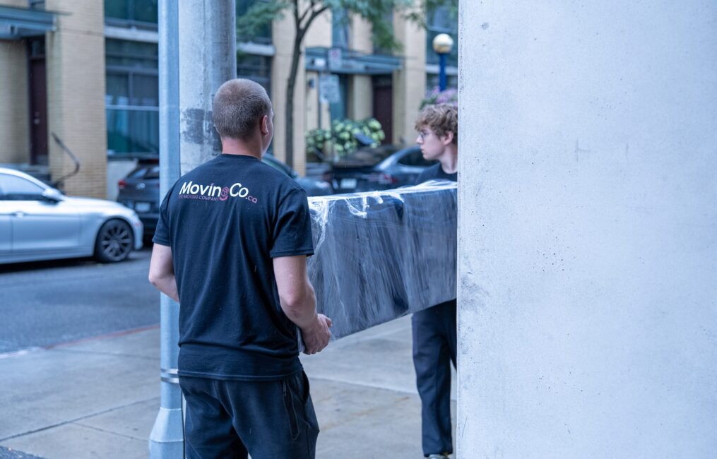 Moving Co. mover carrying a wrapped sofa out of a building with a teammate
