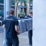 Moving Co. mover carrying a wrapped sofa out of a building with a teammate