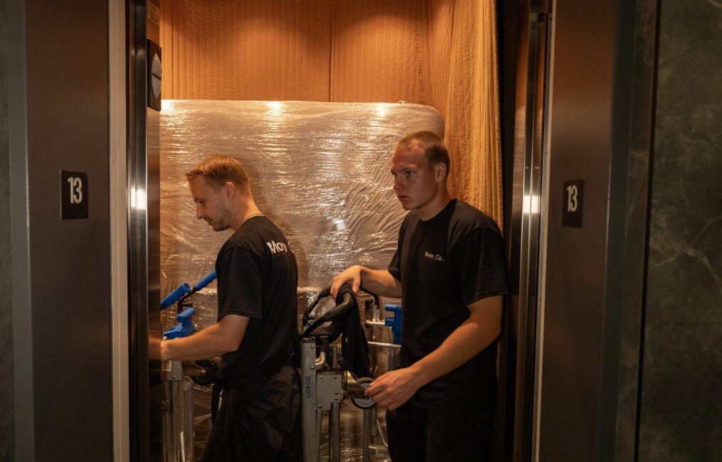 Two Moving Co. movers in a padded condo service elevator with shrink-wrapped furniture