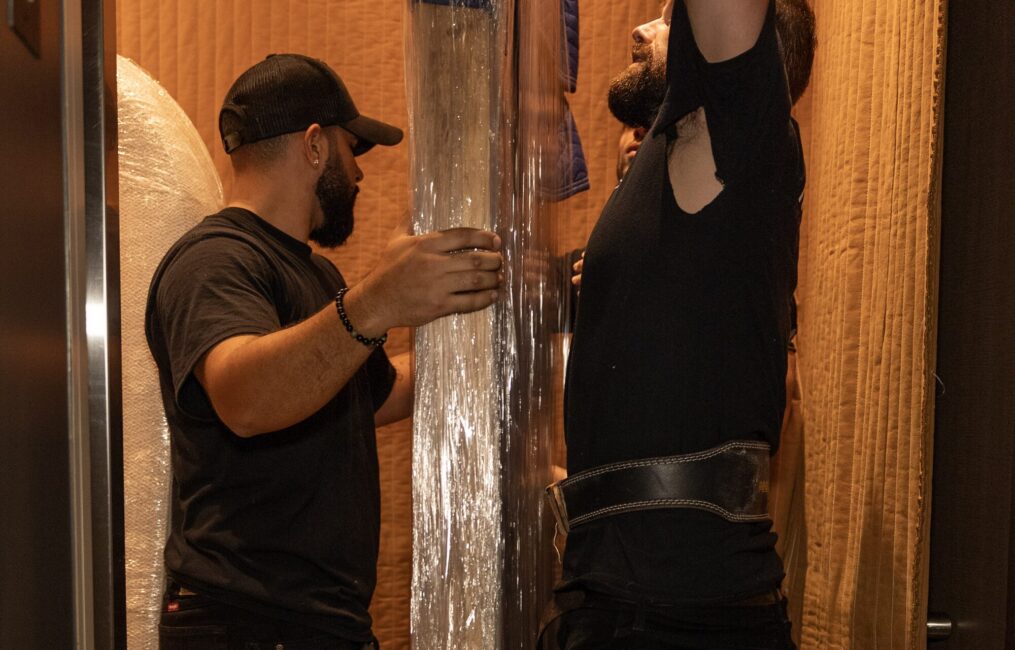 Two Moving Co. movers in a building elevator steadying a tall shrink-wrapped item