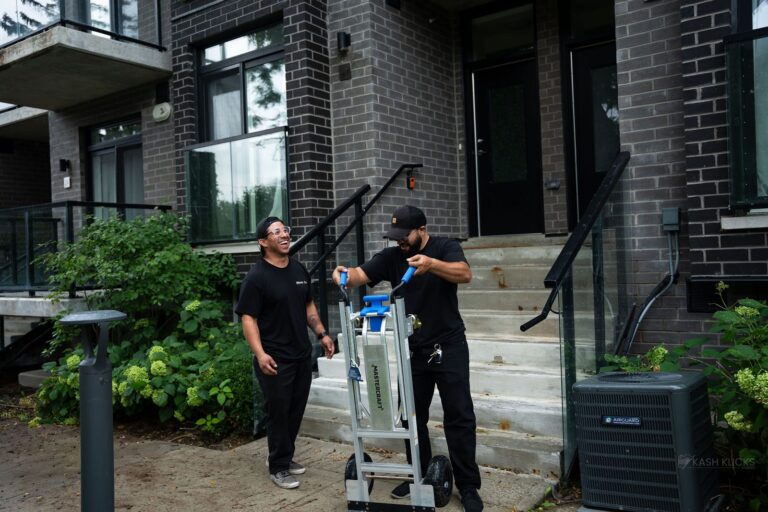 Two Moving Co. movers with a hand truck at the front steps of a townhouse