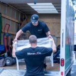 Two Moving Co. movers passing a white shelving unit into the truck