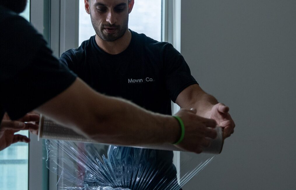 Two Moving Co. movers shrink-wrapping a long piece of furniture beside a condo window