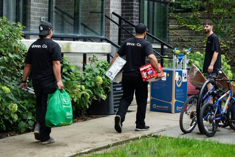 Three Moving Co. movers carrying boxes and bags toward a row of townhouses