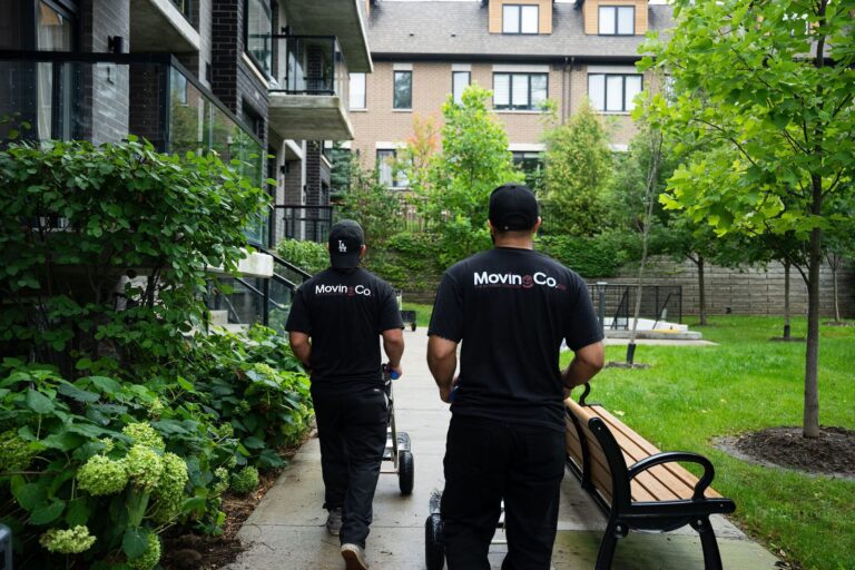 Two Moving Co. movers wheeling hand trucks past a bench in a townhouse complex