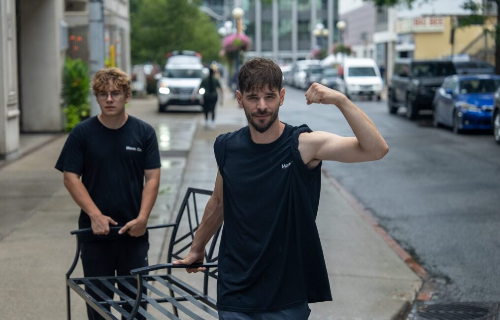 Two Moving Co. movers on the sidewalk with a dolly, one flexing for the camera